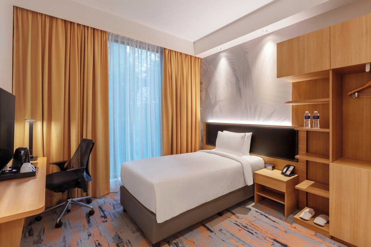 Habitación de hotel con cama, escritorio, silla, televisor, mesita de noche con teléfono y lámpara, armario y ventana con cortinas.