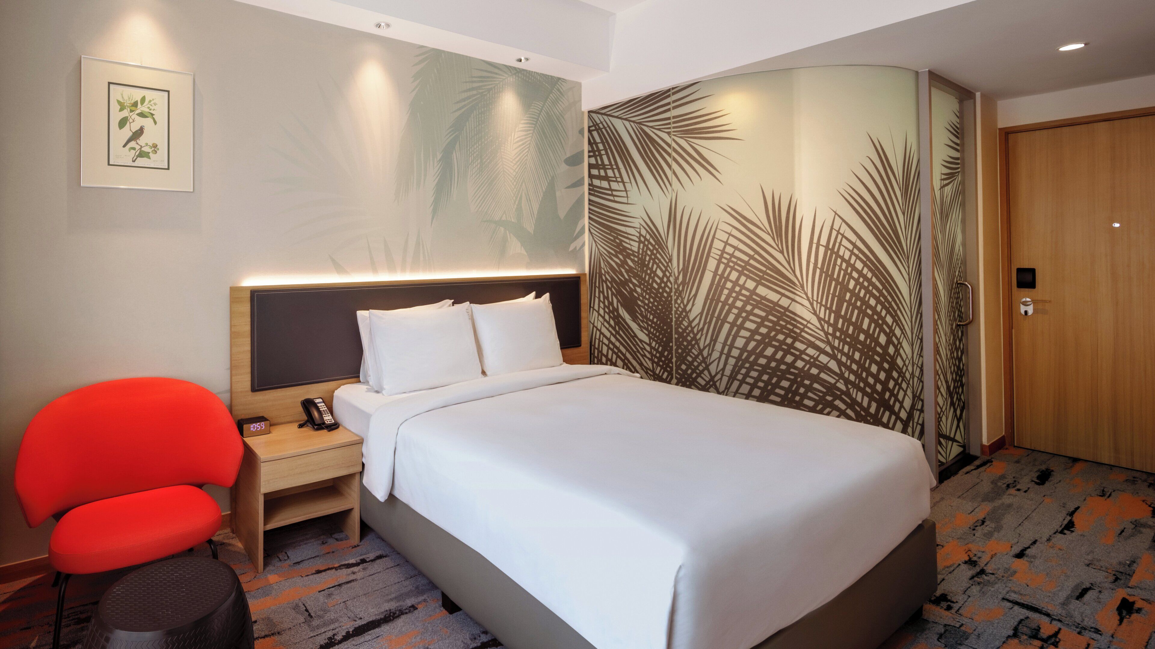 A hotel room with a bed, a nightstand with a phone, a red chair, and a wall with palm leaf patterns.