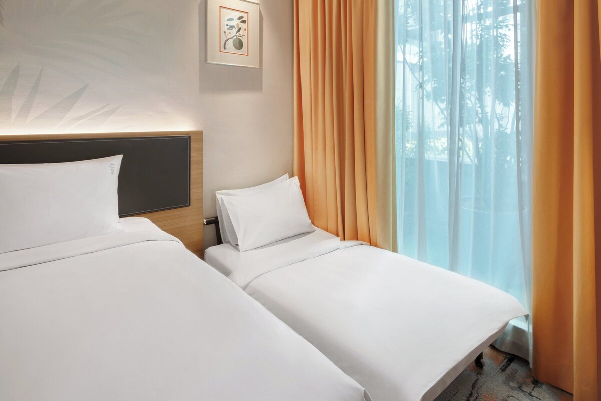 Habitación de hotel con dos camas individuales, cabecera, un cuadro enmarcado en la pared y una ventana con cortinas.