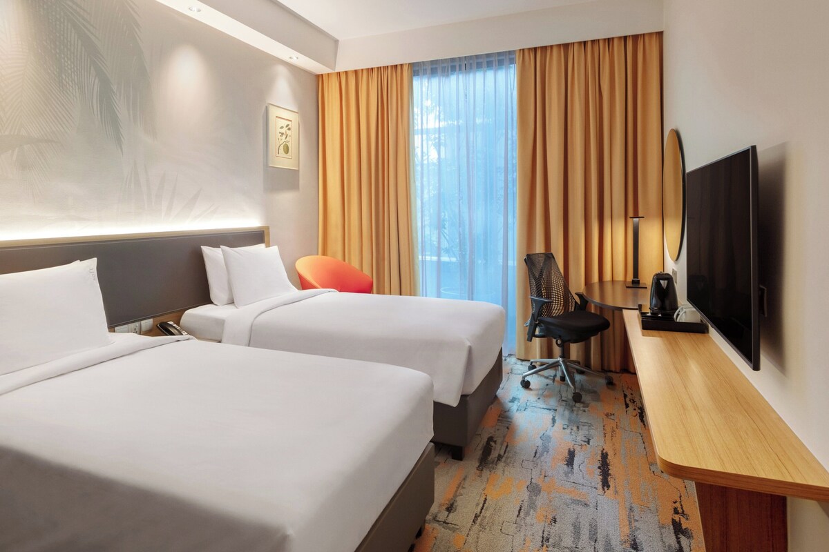 Habitación de hotel con dos camas, un escritorio, una silla, un televisor y un ventanal con cortinas.