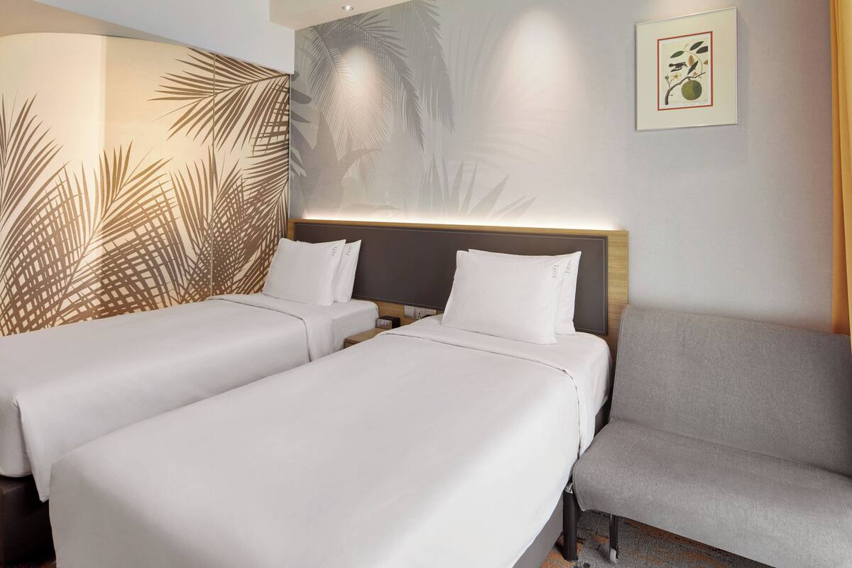 A hotel room with two single beds, a grey armchair, and a decorative wall with palm leaf patterns.