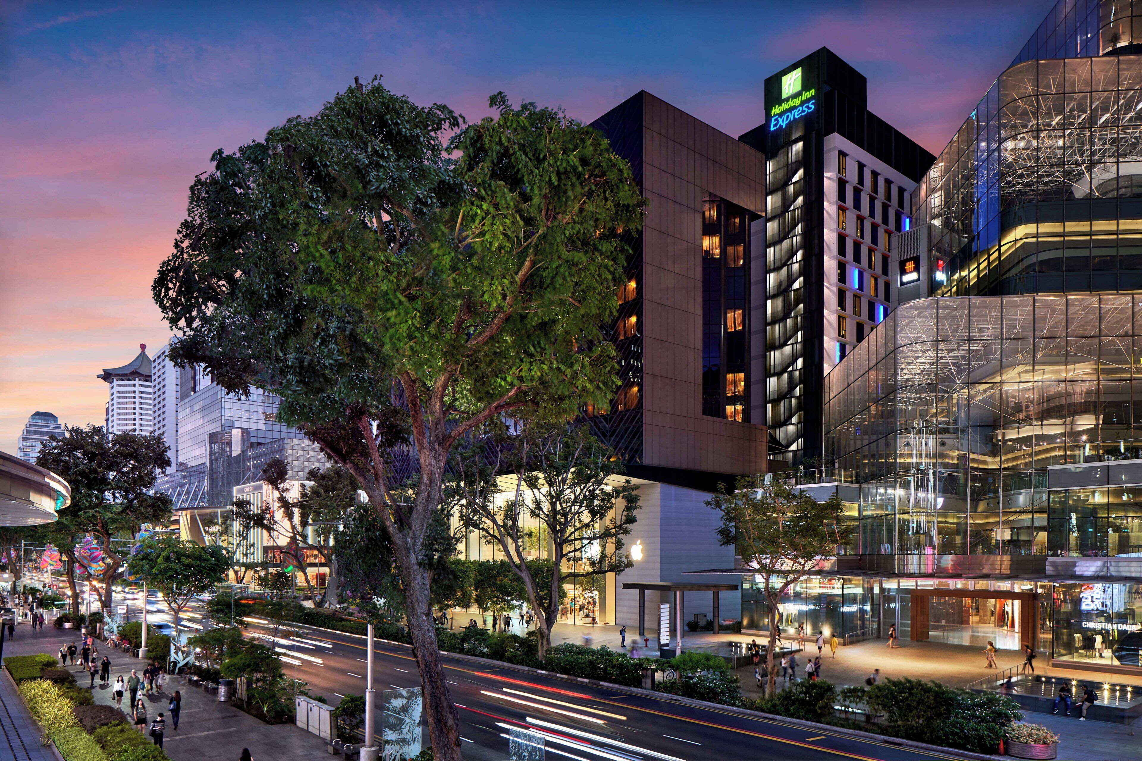 A modern hotel with a prominent Holiday Inn Express sign, surrounded by urban buildings and a busy street at dusk.