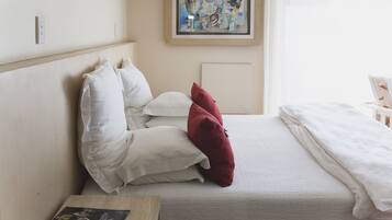 Una cama bien hecha con almohadas blancas y rojas, un cuadro abstracto enmarcado en la pared y una mesita de noche con un libro.