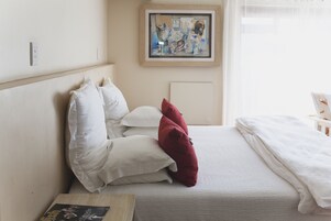 Una cama bien hecha con almohadas blancas y rojas, un cuadro abstracto enmarcado en la pared y una mesita de noche con un libro.