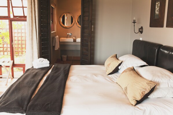 A hotel room with a bed, a bathroom with a sink and mirrors, and a view of the outdoors. - Dolphin Dunes Guest House (Wilderness)