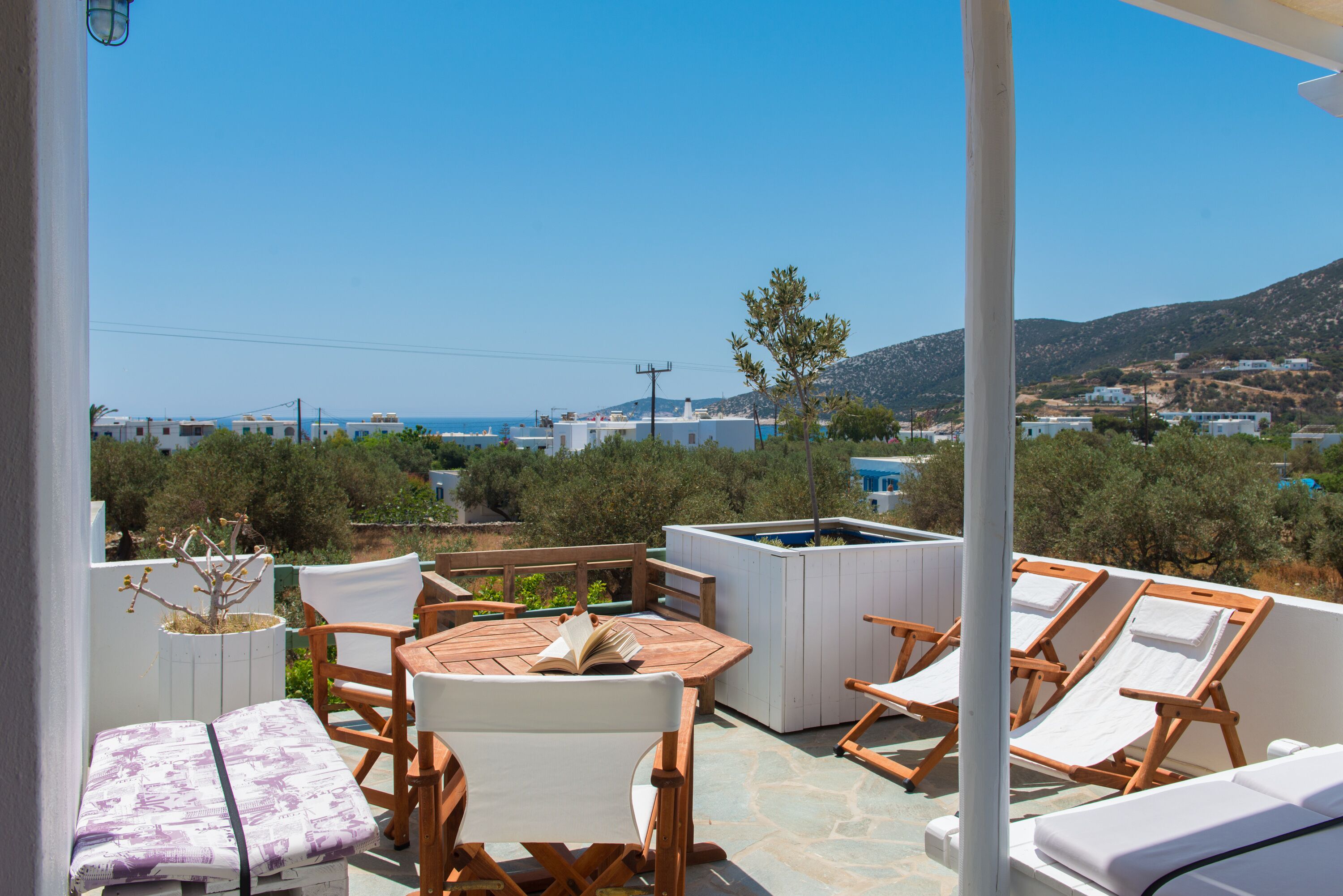 A terrace with wooden furniture, a table, and a view of the sea and hills.