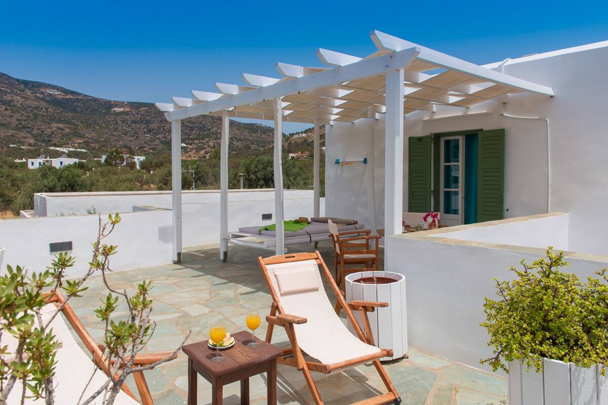 A rooftop terrace with a pergola, lounge chairs, a table with drinks, and a view of the landscape.