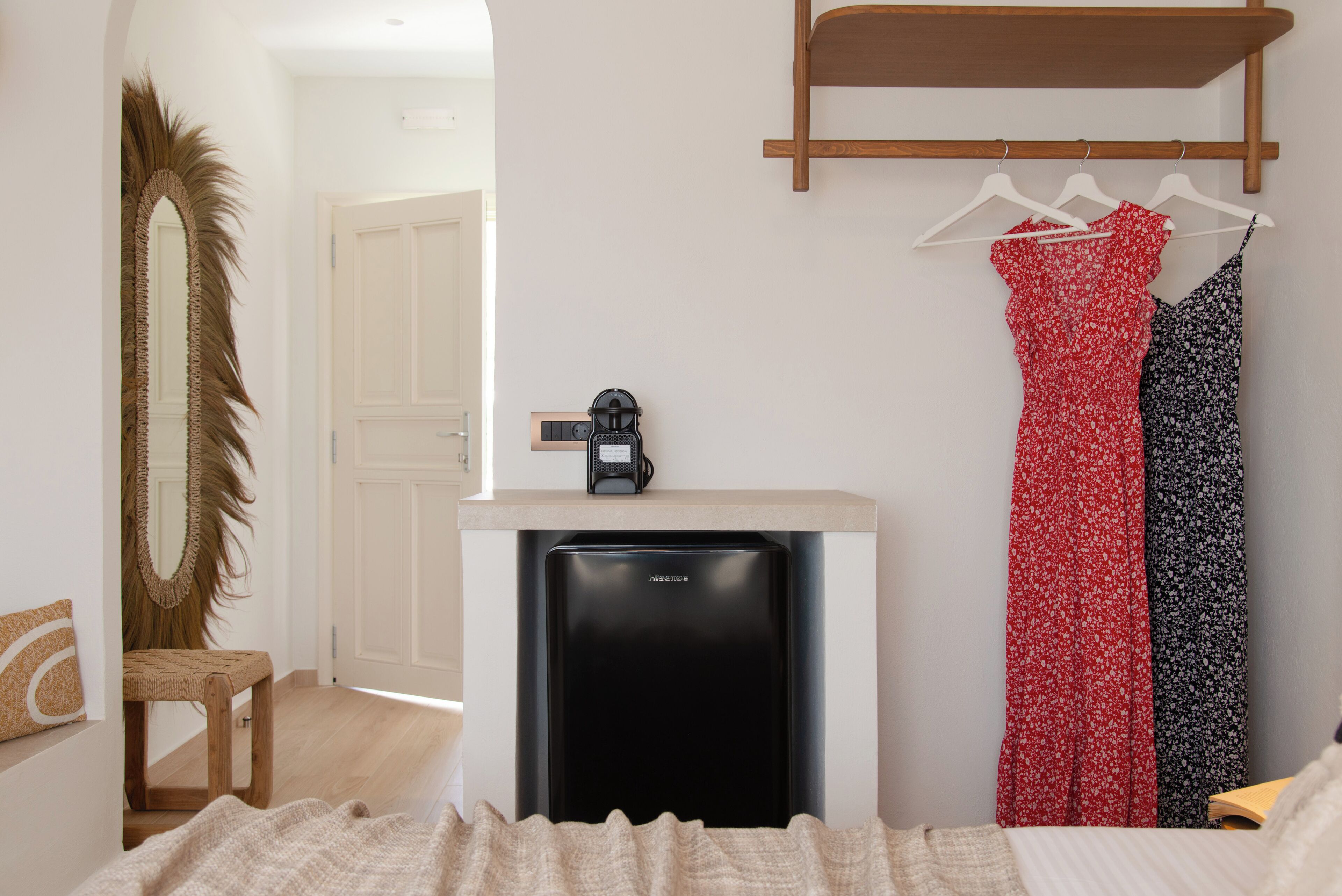 A compact room with a compact refrigerator, a wooden chair, a bed with a beige blanket, and a clothing rack with two dresses.