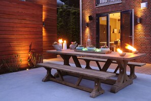 An outdoor wooden picnic table with candles and flowers, set against a brick building with a sliding glass door.