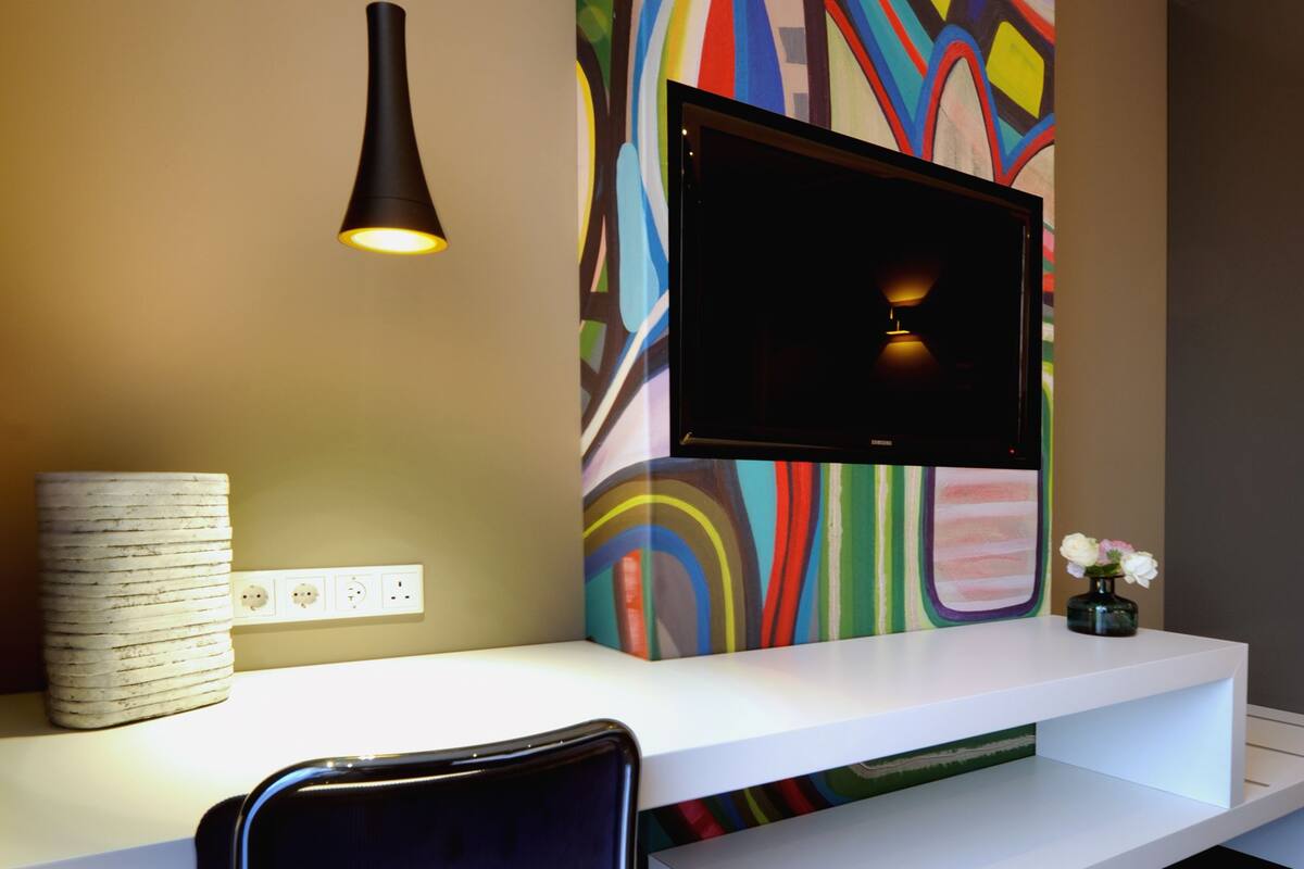 Een moderne receptiebalie met een flatscreen televisie bevestigd aan een kleurrijke, abstracte muur, een witte console tafel, een zwarte stoel en een hanglamp.