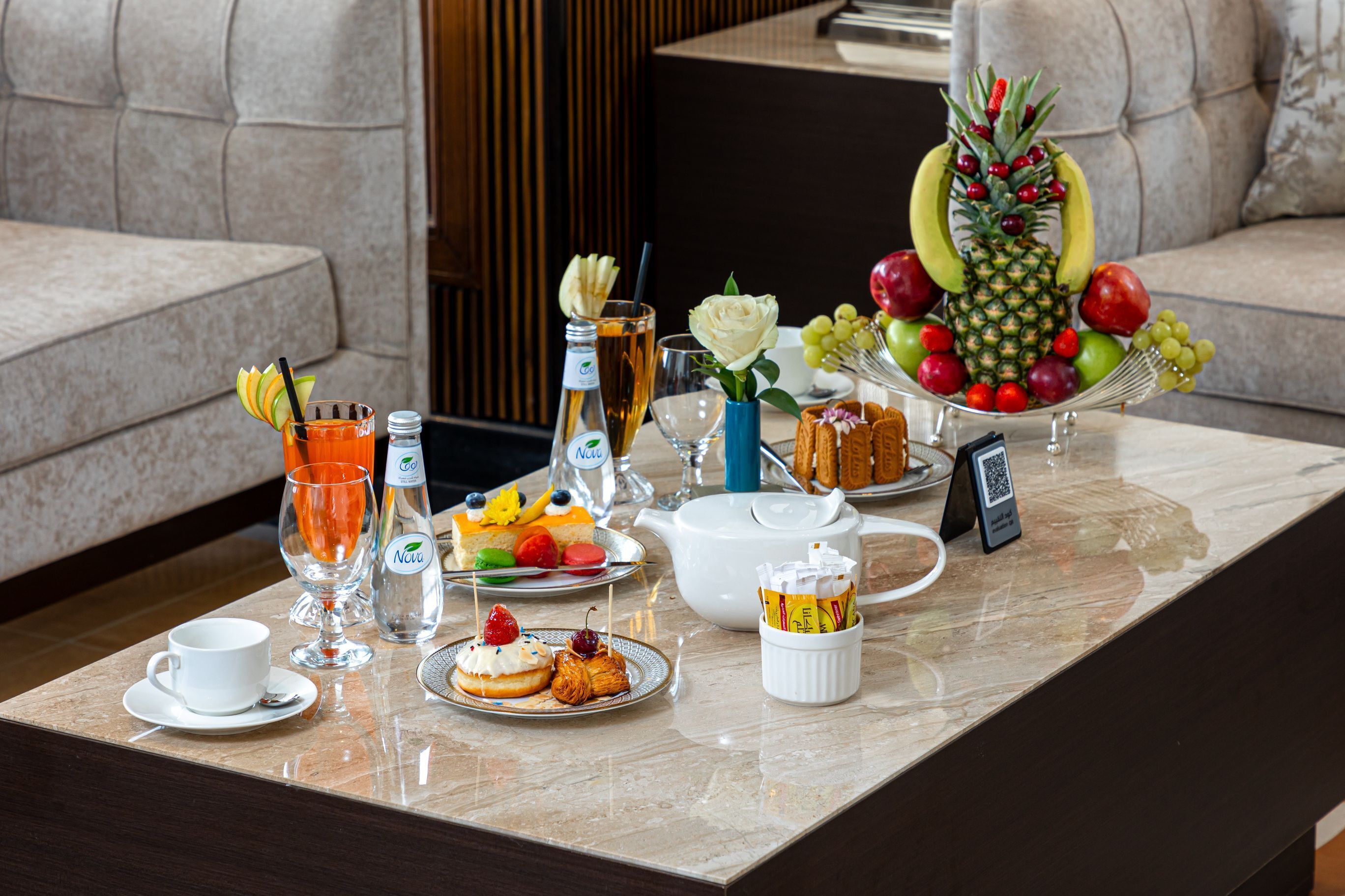 A breakfast setup with a variety of fruits, pastries, and beverages on a marble table in a modern living room with a sofa.