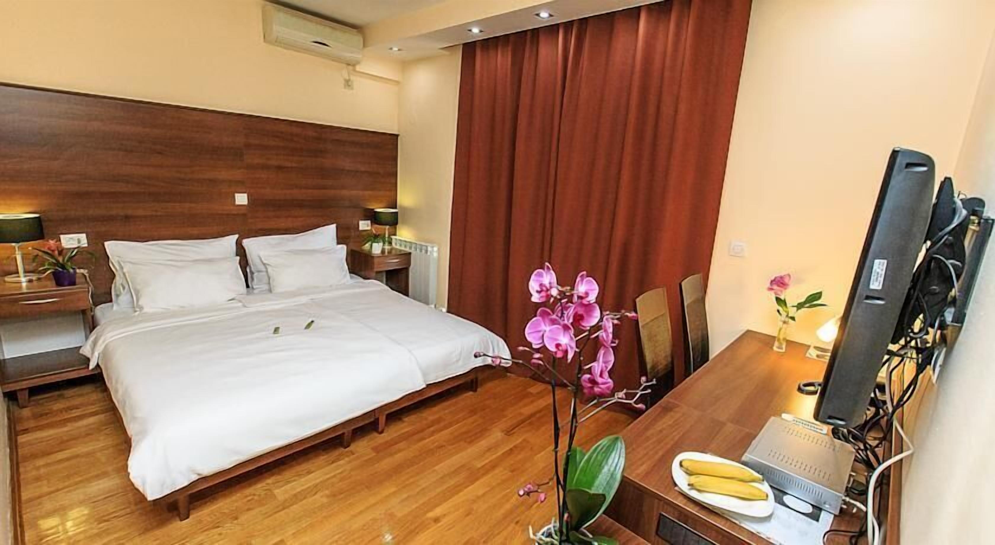 A hotel room with a large bed, wooden headboard, two bedside tables with lamps, a desk with a computer and banana, and a large window with curtains.