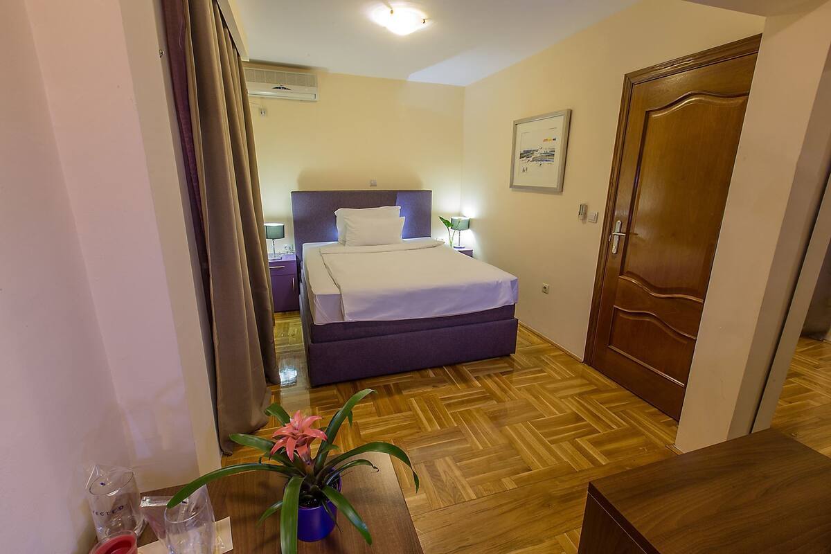 A hotel room with a bed, a wooden floor, a door, and a wall-mounted air conditioner.