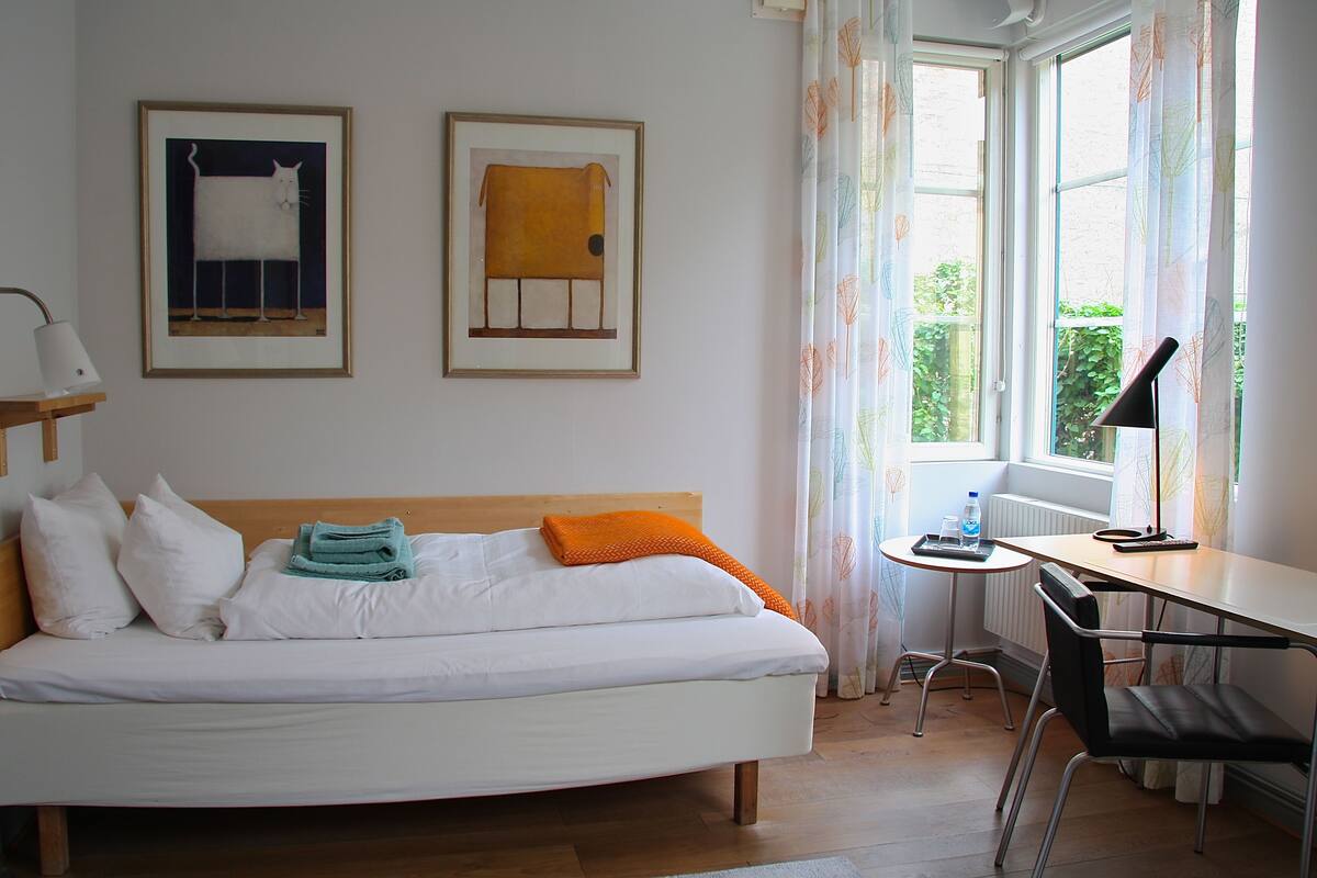 A hotel room with a bed, a desk, and two framed artworks on the wall.
