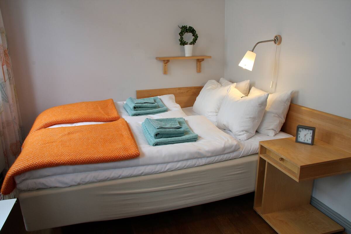 A neatly made bed with white and orange bedding, a small wooden nightstand with a clock, and a wall-mounted shelf with a plant.