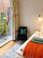 Una habitación con cama, una silla, ventana con cortinas, lámpara y vista a un edificio de ladrillo y árboles.