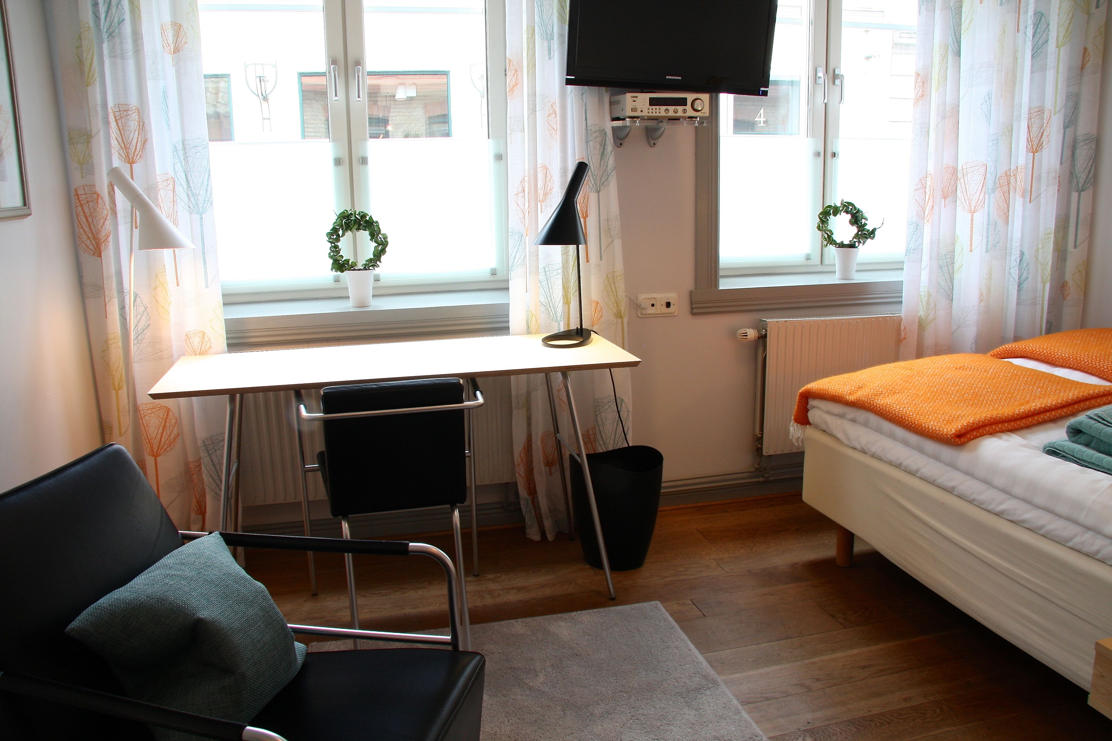 A bedroom with a bed, a desk, a chair, a TV, and a window with curtains.