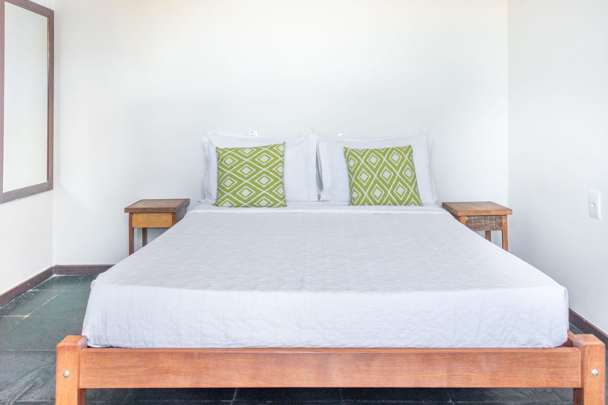 A single bed with a white mattress and two pillows, one green and one white, in a room with white walls and a wooden headboard.