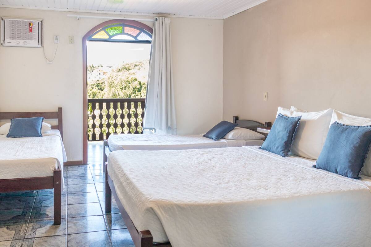 A hotel room with three beds, a balcony with a decorative railing, and a view of greenery outside.