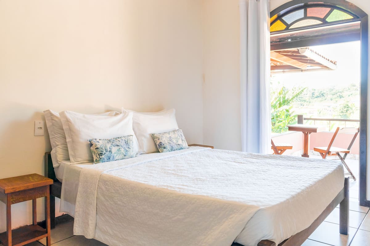 A bed with white bedding and floral pillows, a wooden nightstand, and a view of a balcony with chairs and greenery.