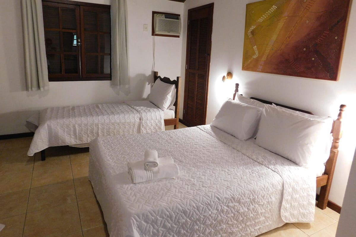 A room with two beds, a window with curtains, an air conditioning unit, and a painting on the wall.
