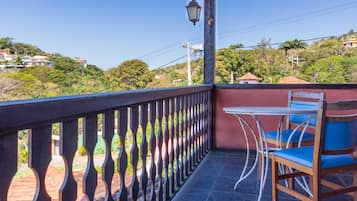 Un balcón con mesa y sillas, barandilla y vistas a una colina con casas y árboles.