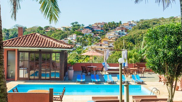 A swimming pool with lounge chairs, a glass-enclosed cabana, and a view of hillside houses. - Pousada dos Coqueiros Búzios (Búzios)