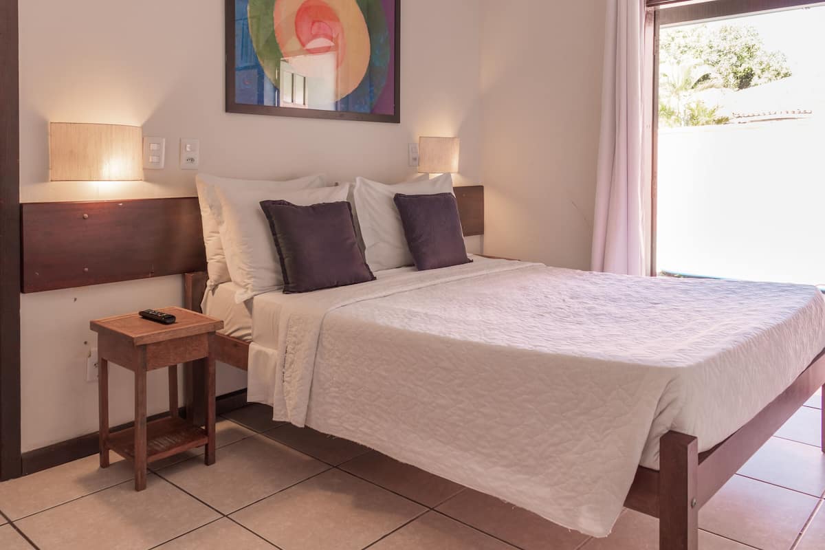 A neatly made bed with white and dark pillows, a wooden bedside table with a remote control, and two wall-mounted lamps.