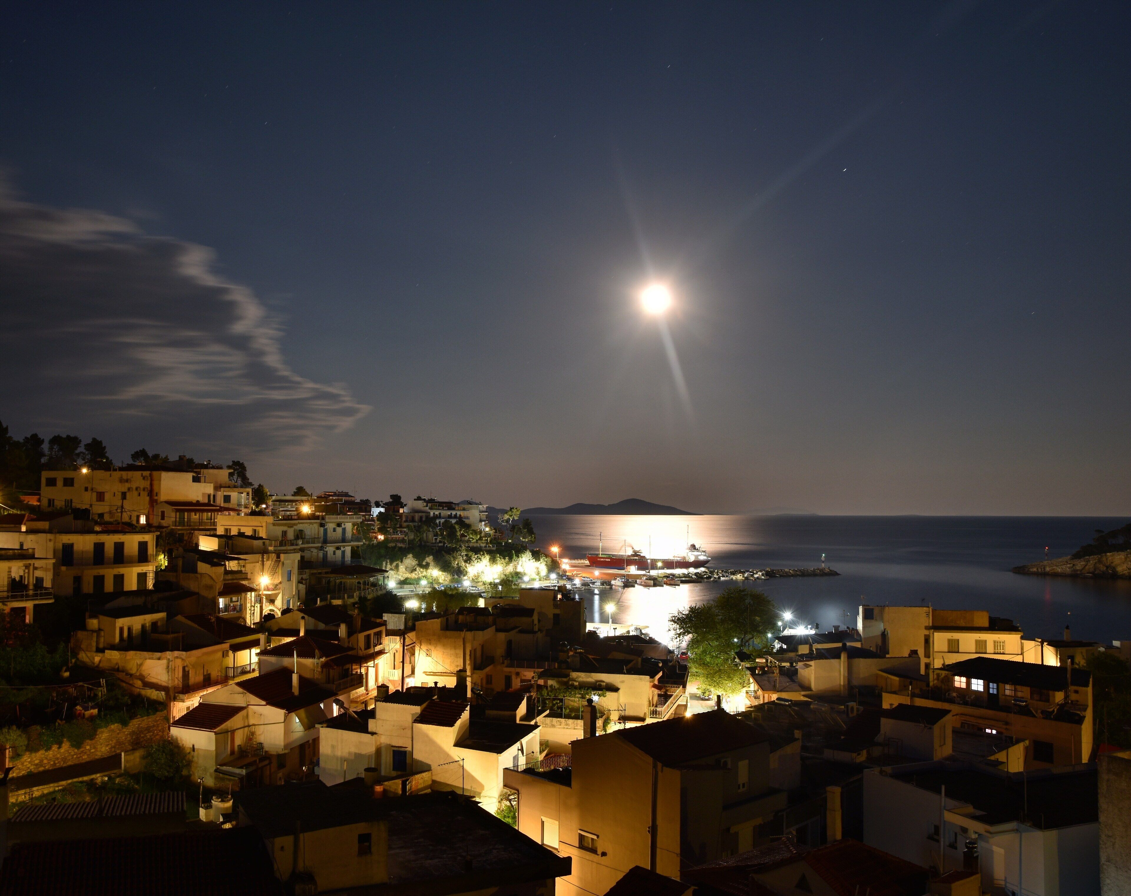 A coastal town at night with buildings illuminated, a boat docked near the shore, and a prominent moon in the sky.