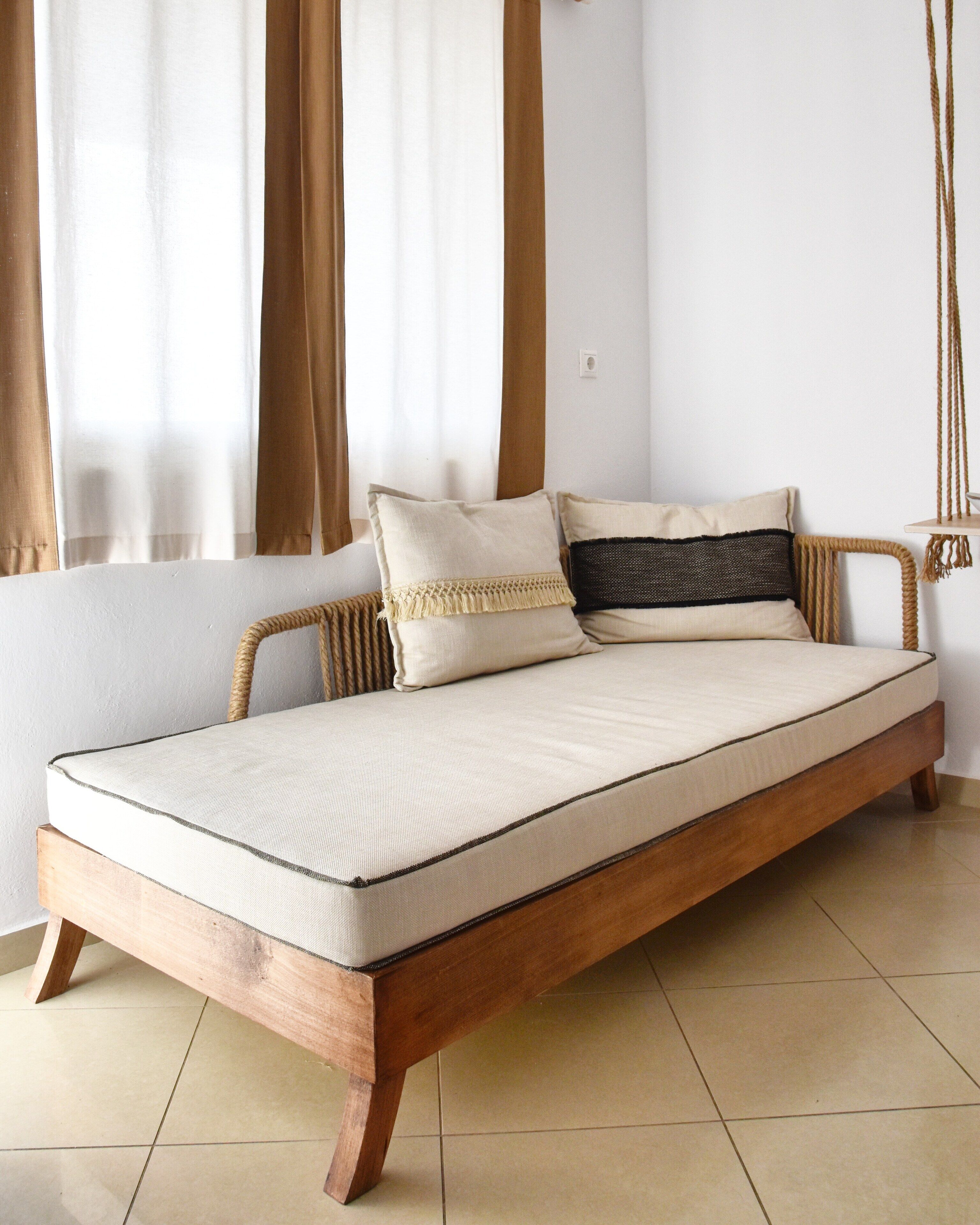 A single bed with a wooden frame and a mattress, positioned in a room with a tiled floor and curtains.