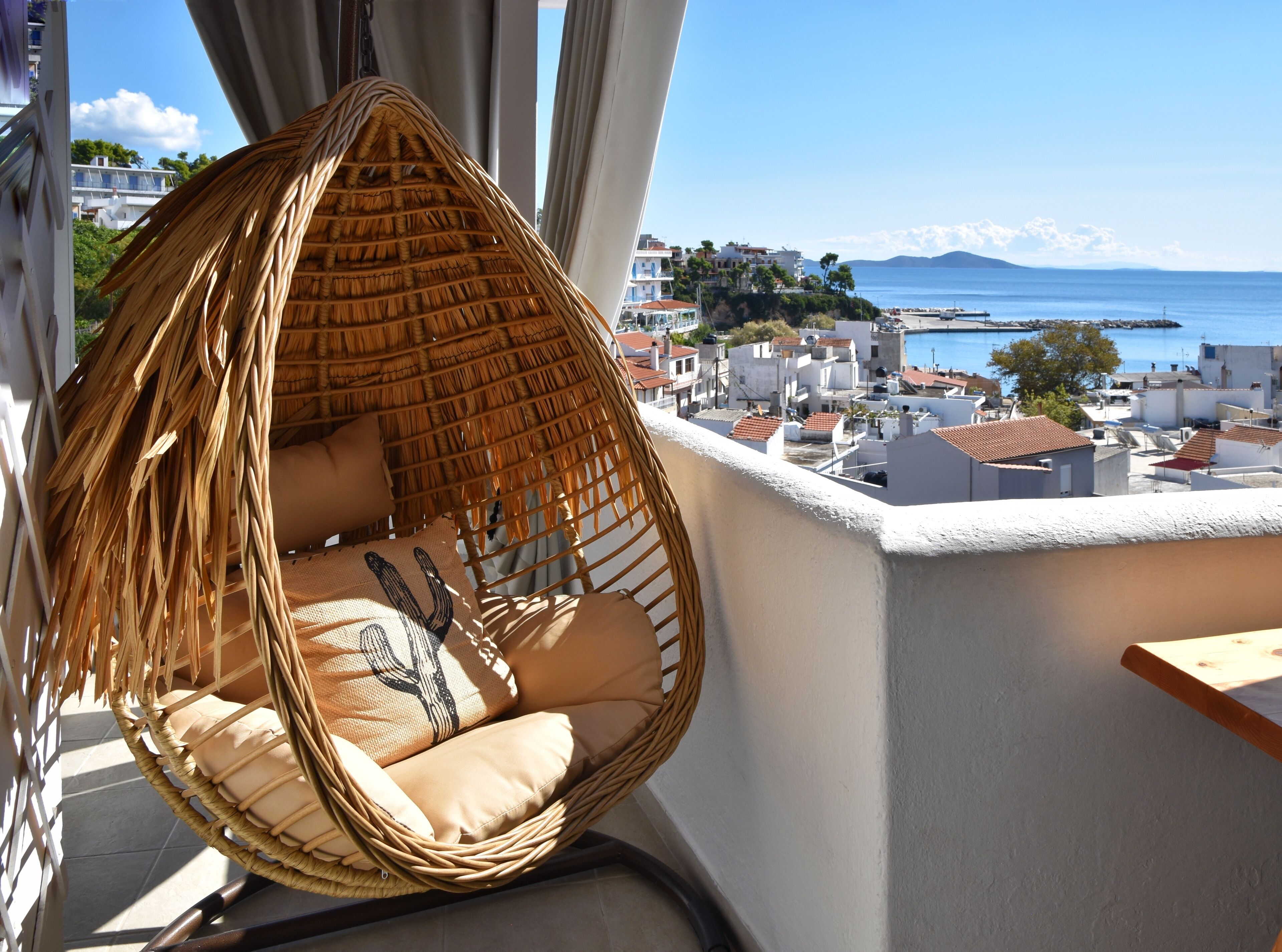A wicker hanging chair with cushions on a balcony overlooking a coastal town with buildings and a clear sky.