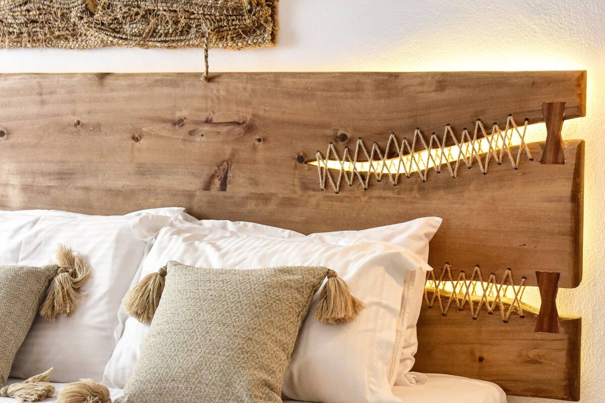 A bed with white linens and two pillows, a wooden headboard with a rope design, and a small round table beside the bed.