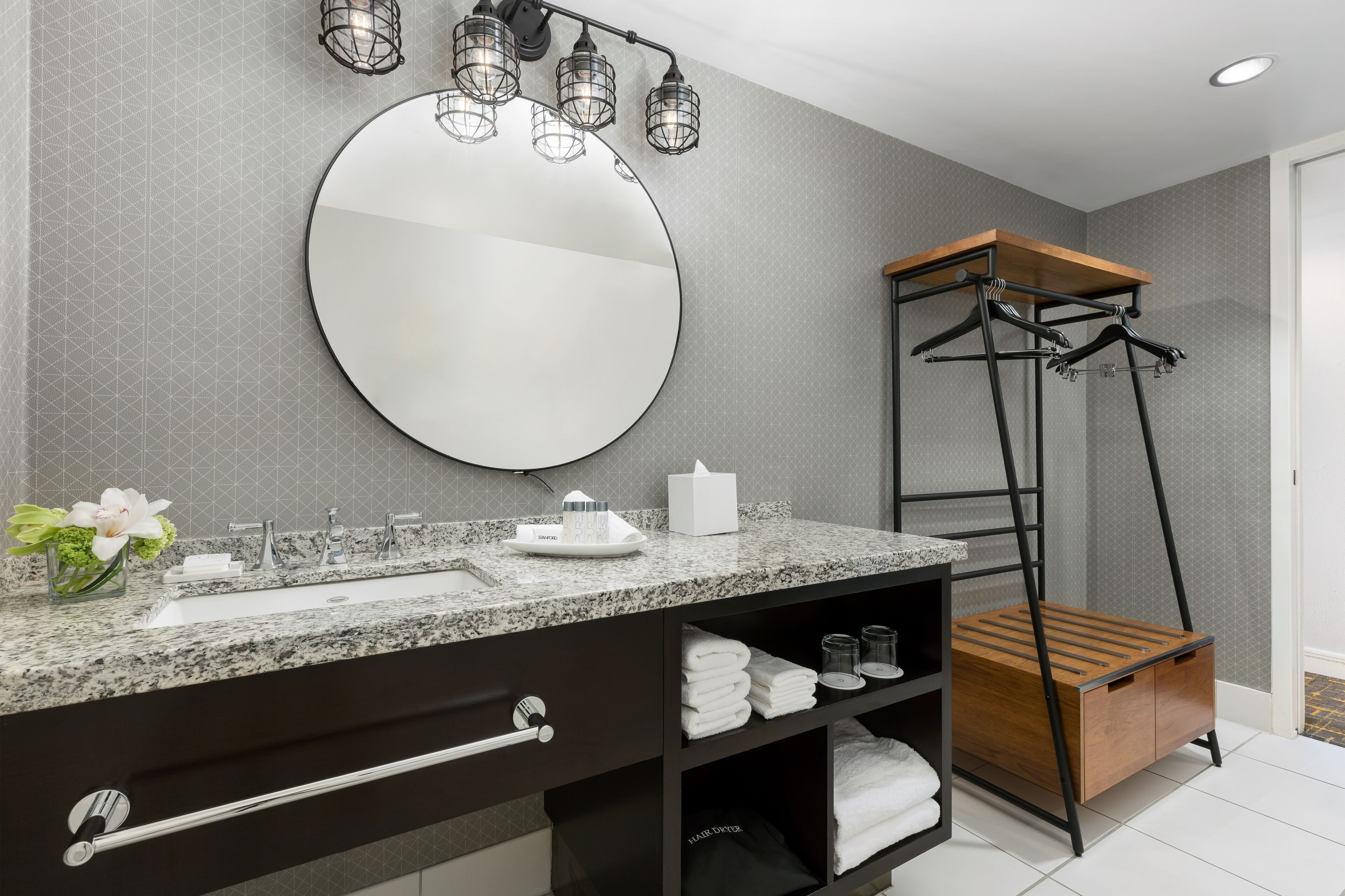 A modern bathroom with a granite countertop, a large mirror, a towel rack, and a wooden shelf unit.