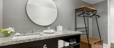A modern bathroom with a granite countertop, a large mirror, a towel rack, and a wooden shelf unit.