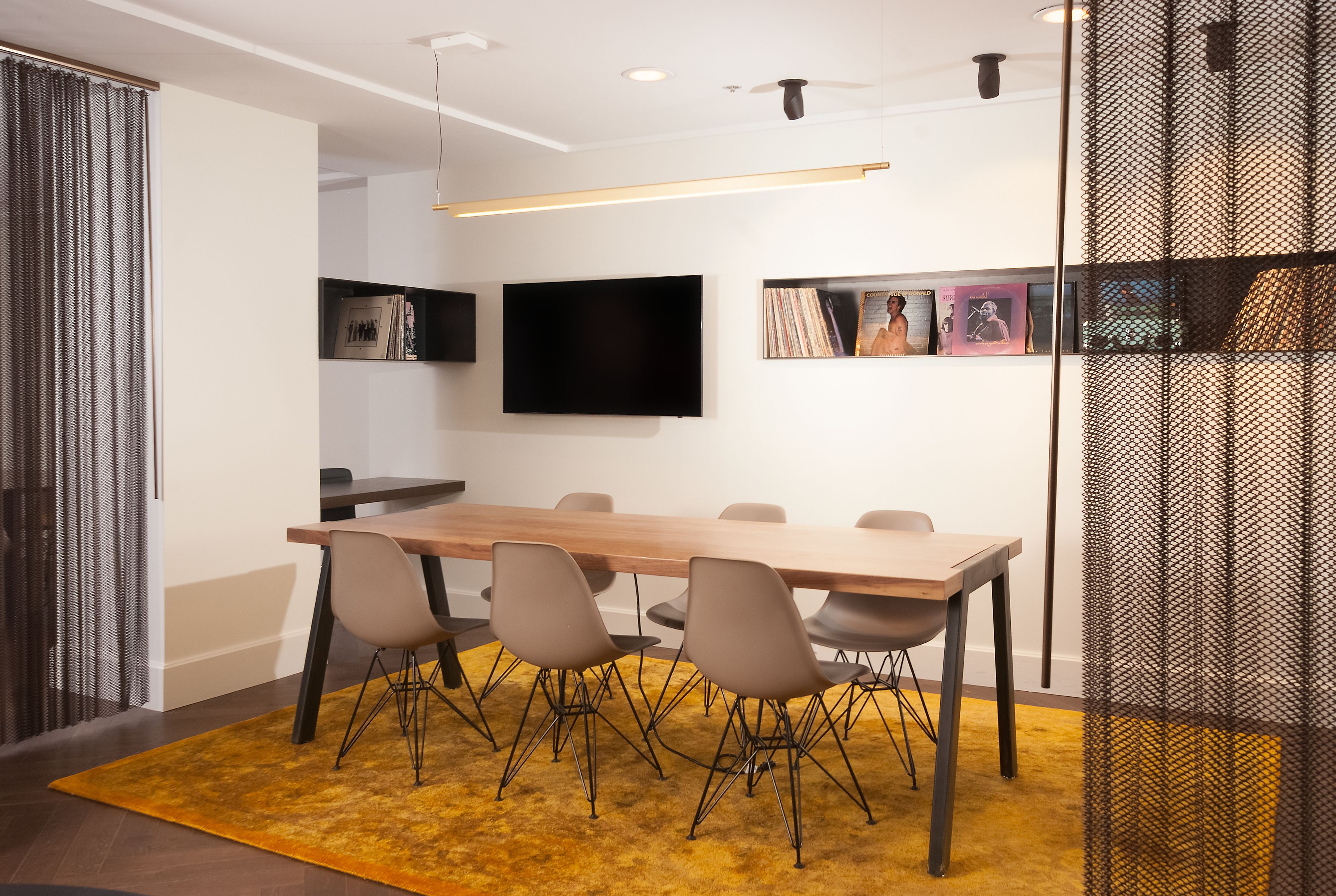 A modern conference room with a long table, six chairs, a flat-screen TV, and a bookshelf with books and framed pictures.