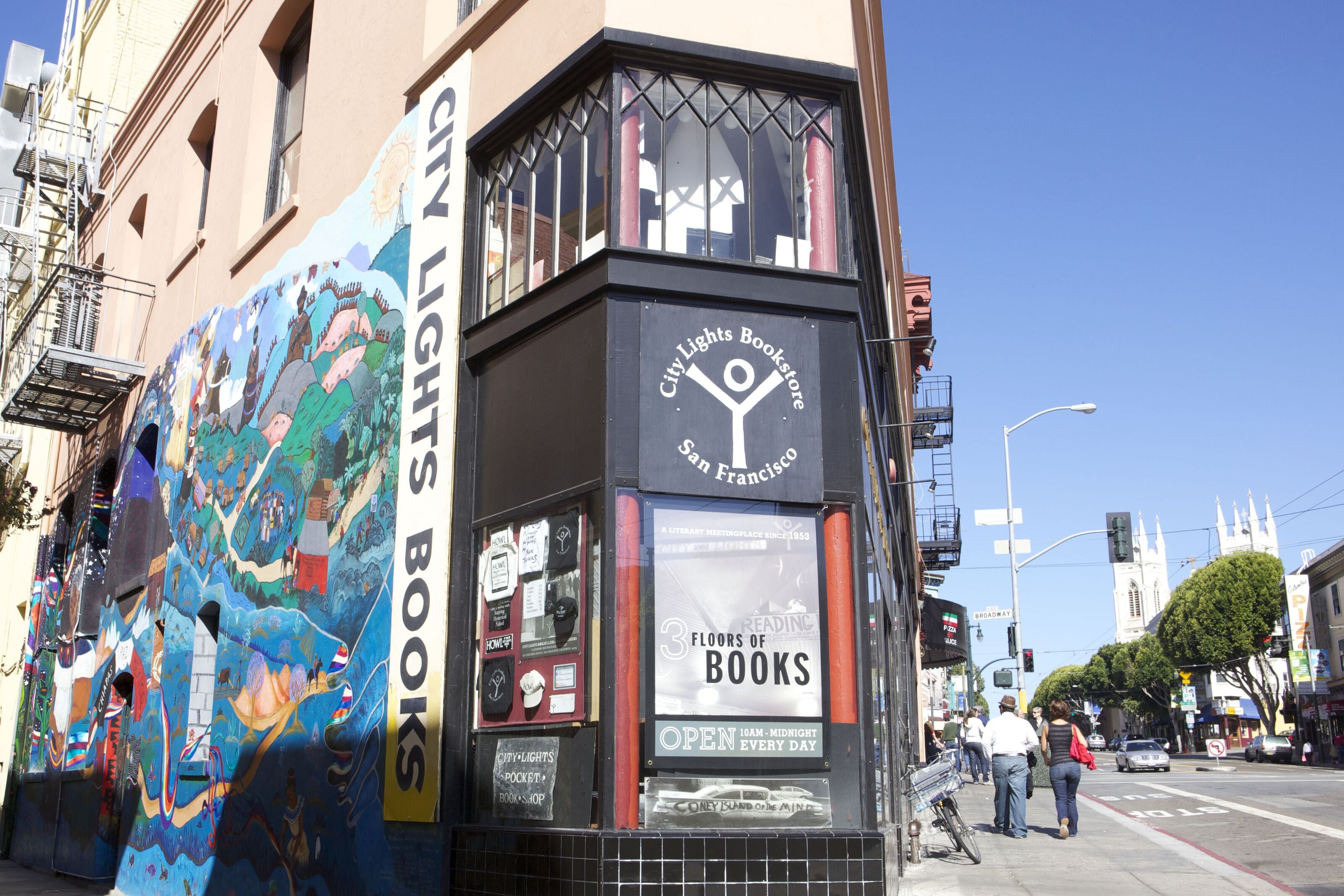 Una librería con un mural colorido en la fachada, un letrero que indica que abre de 10 de la mañana a medianoche todos los días, y una vista de la calle con gente caminando.