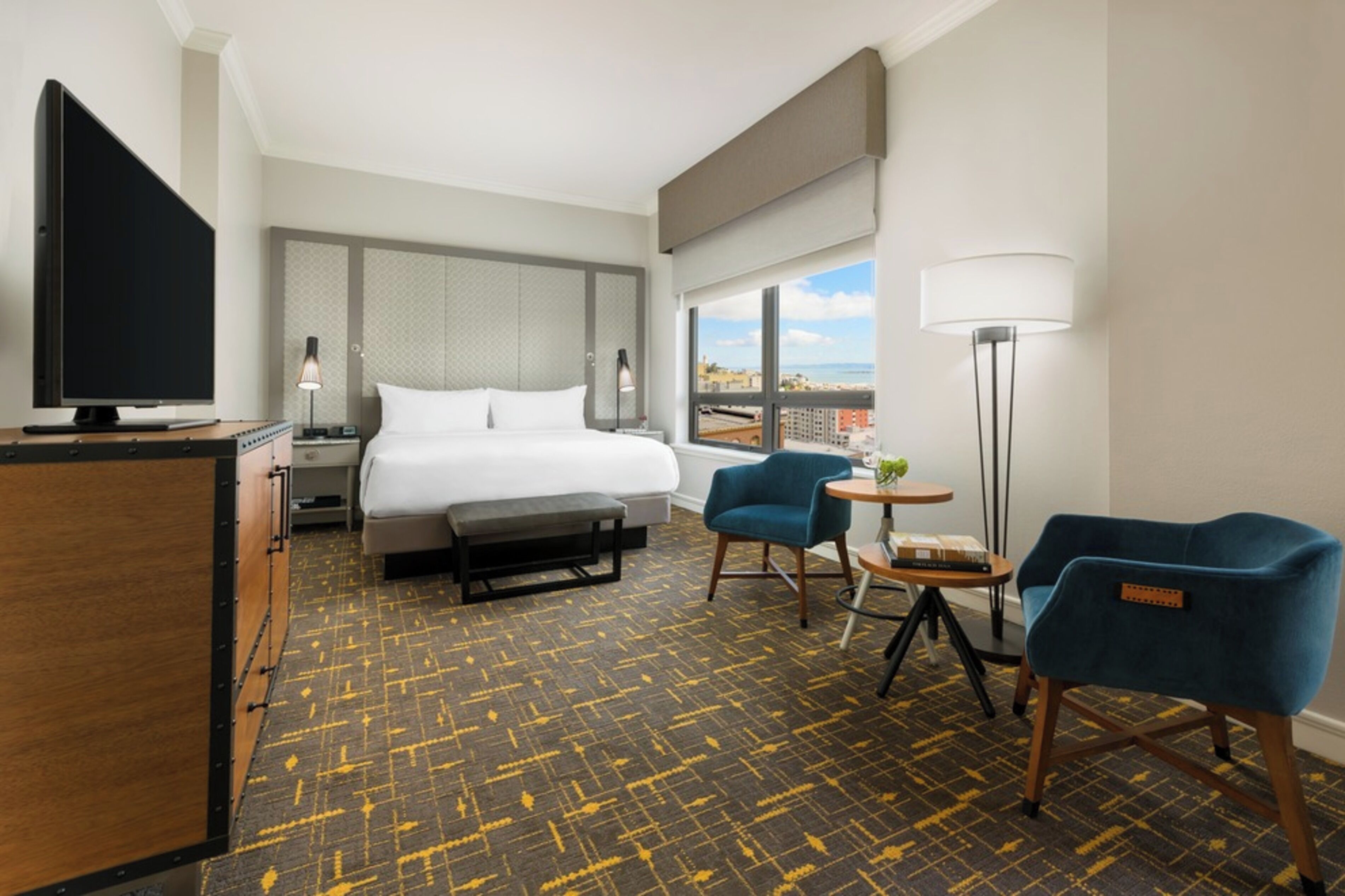 A hotel room with a bed, a desk, a TV, two chairs, a lamp, and a view of the city.