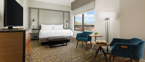 A hotel room with a bed, a desk, a TV, two chairs, a lamp, and a view of the city.