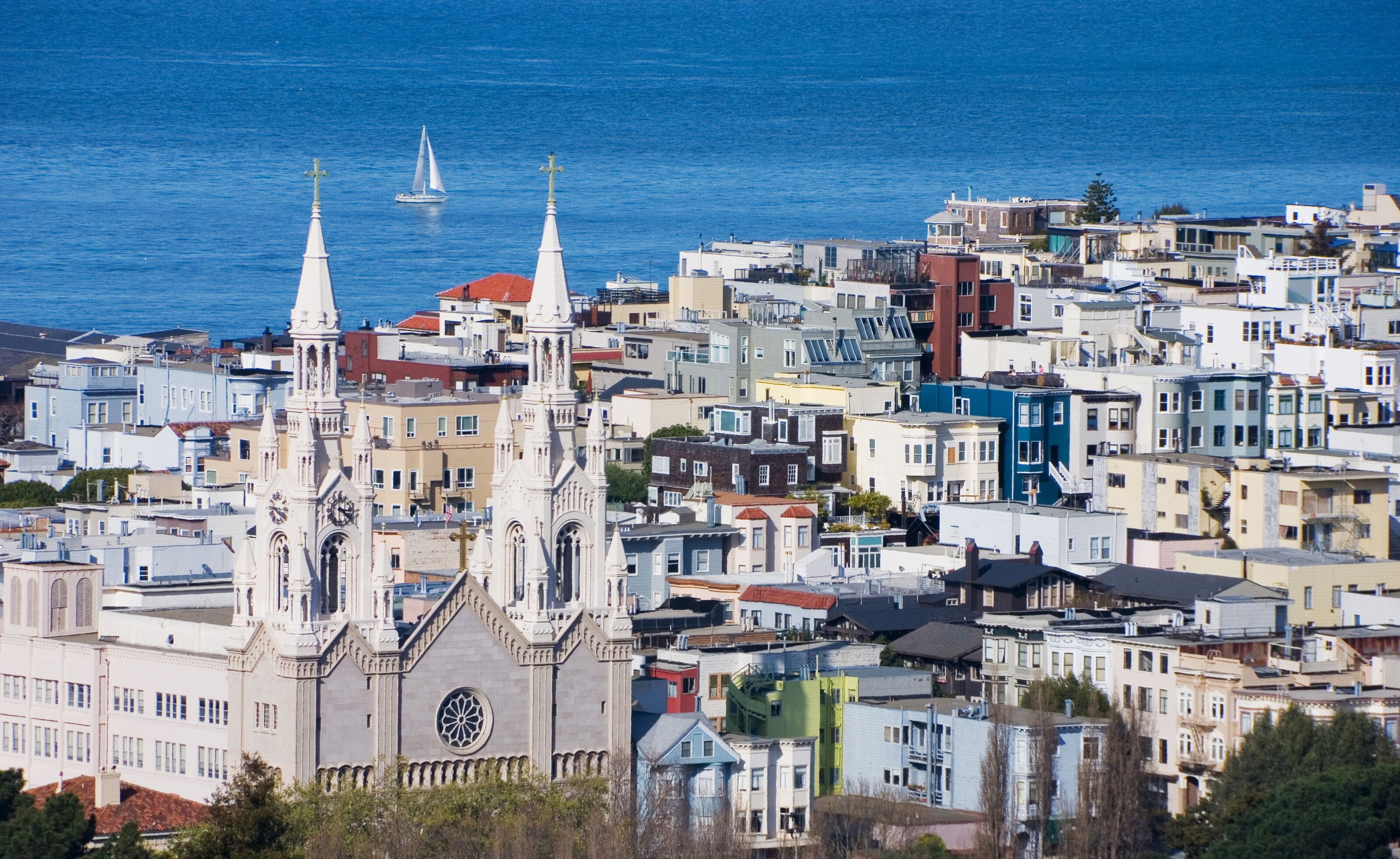 A historic church with two spires and a large circular window, surrounded by densely packed urban buildings and a sailboat on the water.