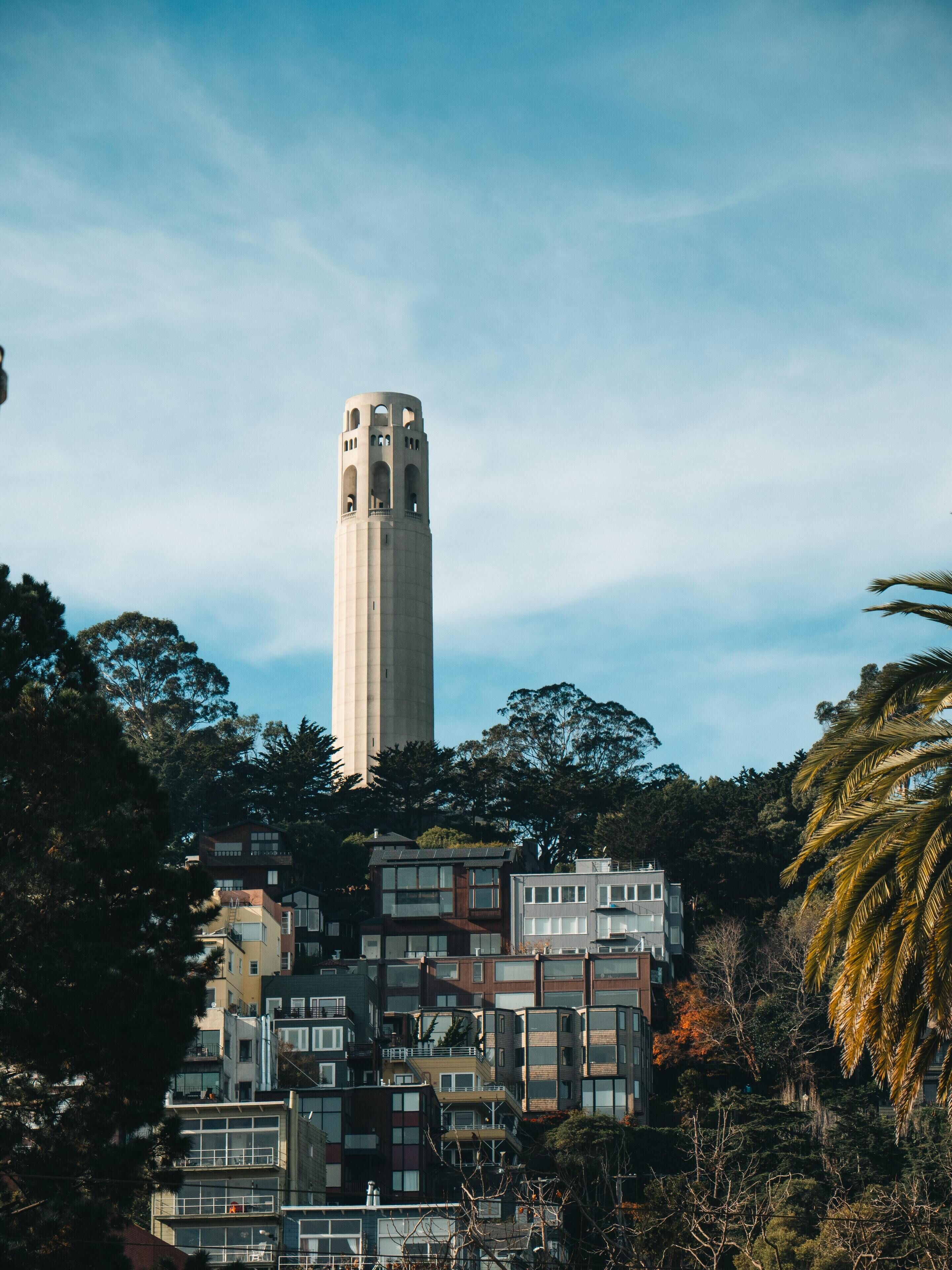 Una torre alta con varios balcones, rodeada de árboles frondosos y edificios modernos.