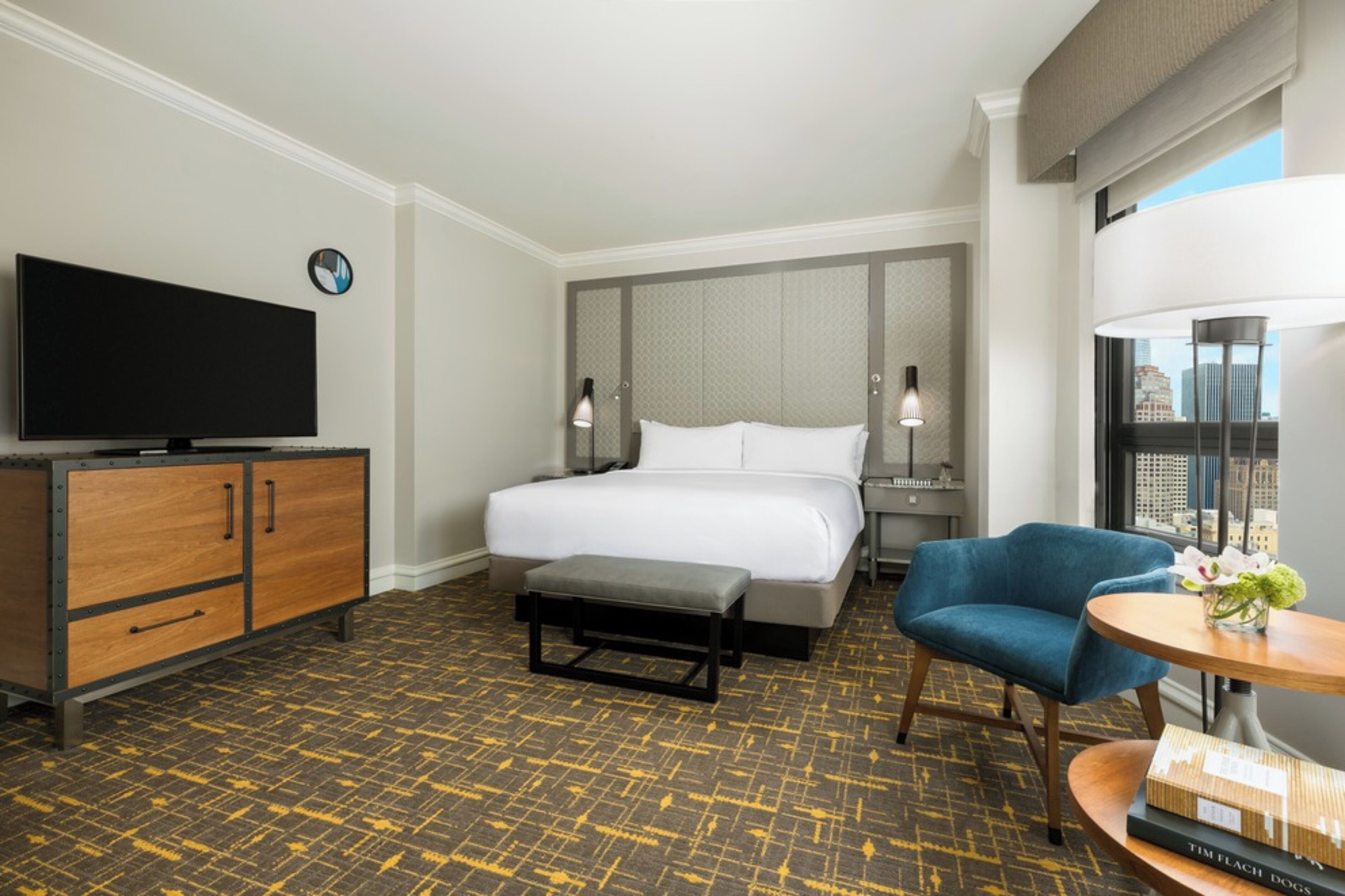 A hotel room with a bed, a television, a desk with a lamp, a chair, and a view of the city.