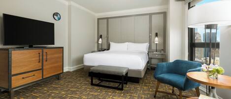 A hotel room with a bed, a television, a desk with a lamp, a chair, and a view of the city.