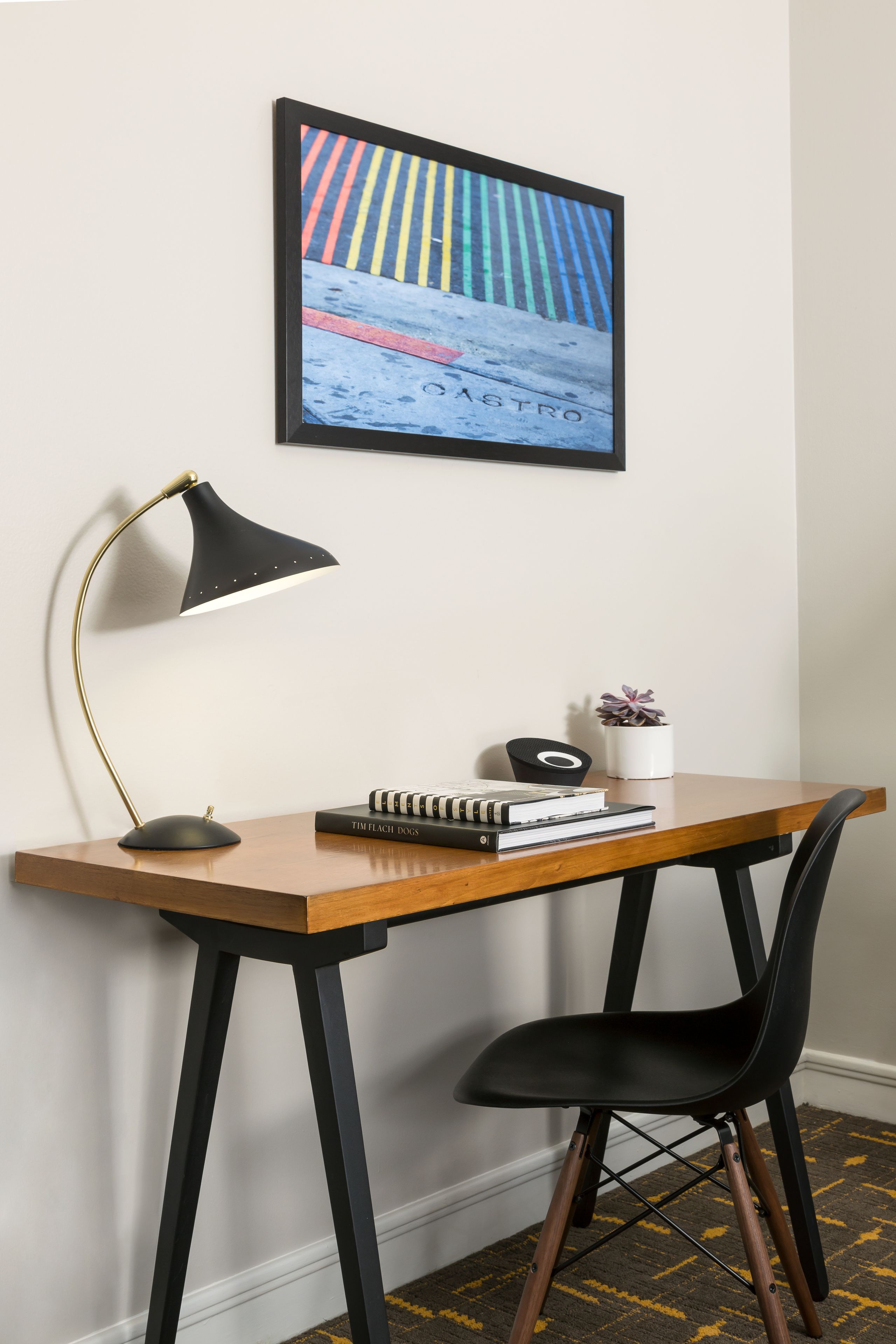A wooden desk with a black chair, a black desk lamp, a framed picture, and a small potted plant.