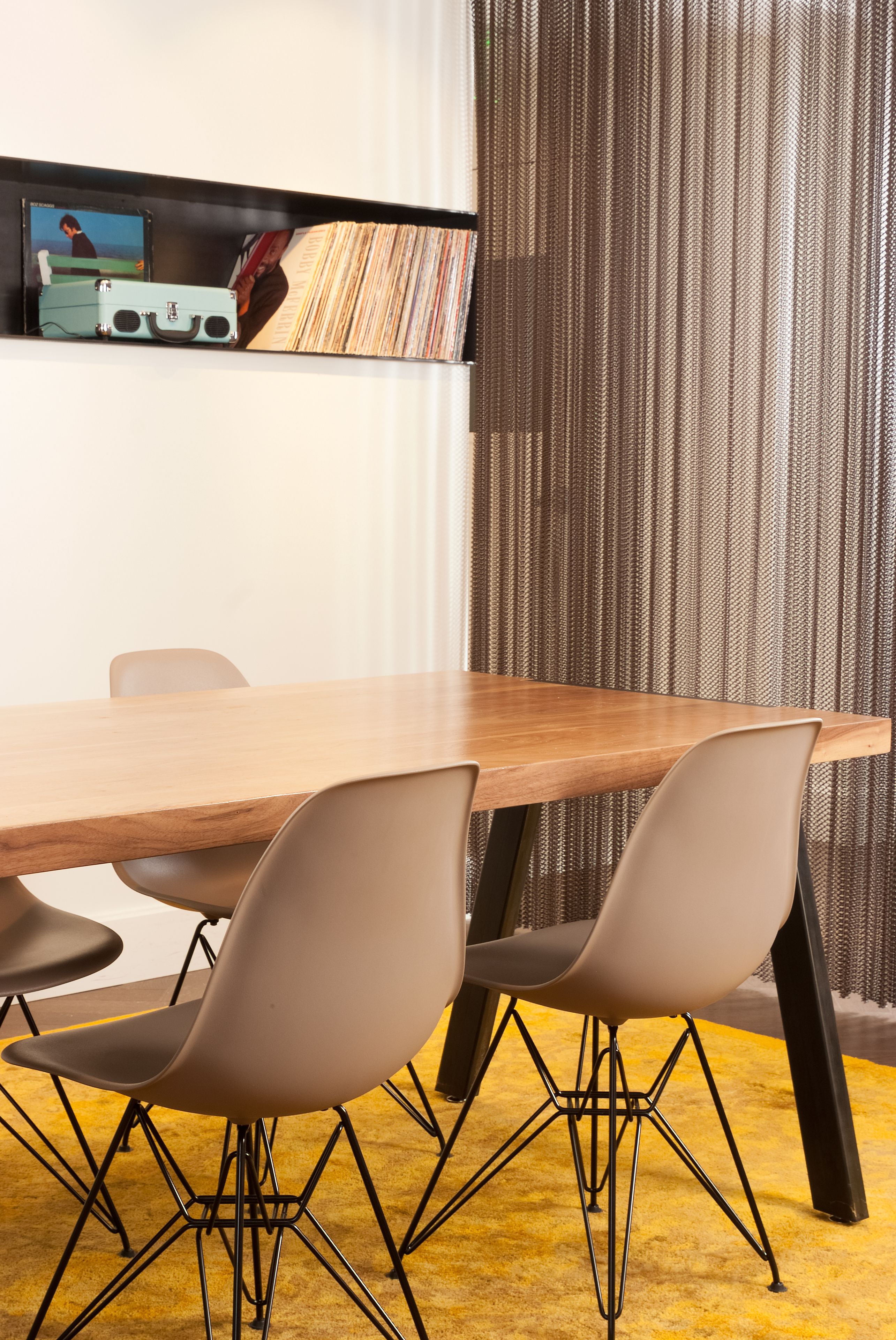 A conference room with a wooden table, four chairs, a shelf with books and a suitcase, and a curtain backdrop.
