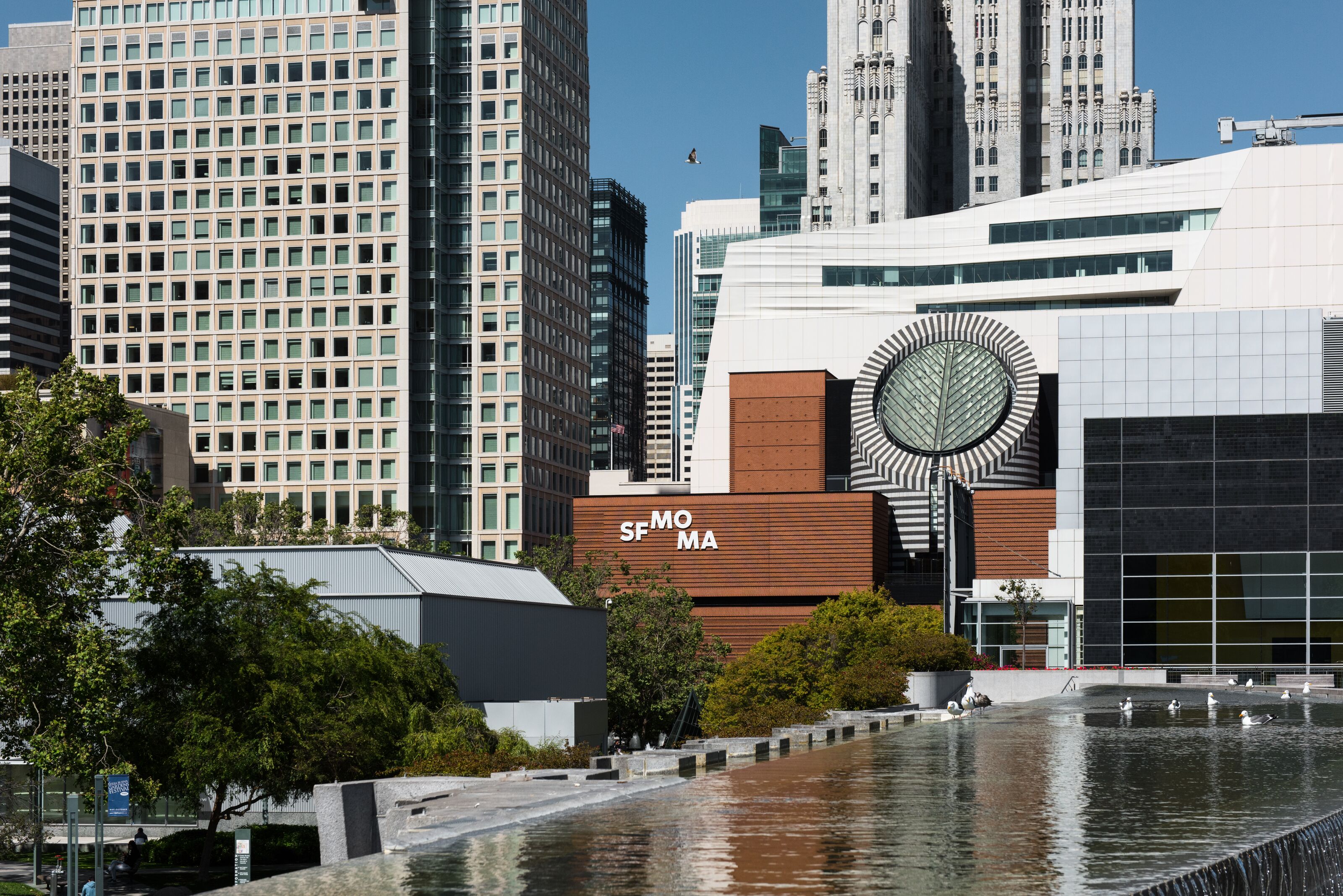 A modern building with a distinctive circular design element, surrounded by a water feature and urban skyscrapers.