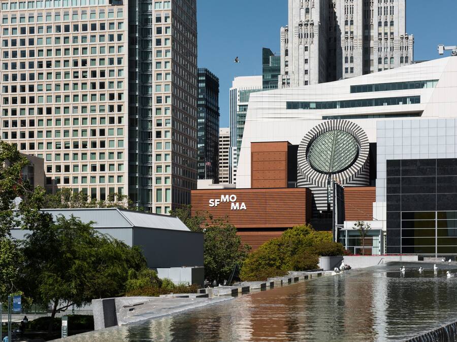 Um edifício moderno com um elemento de design circular marcante, rodeado por um espelho d'água e arranha-céus urbanos.