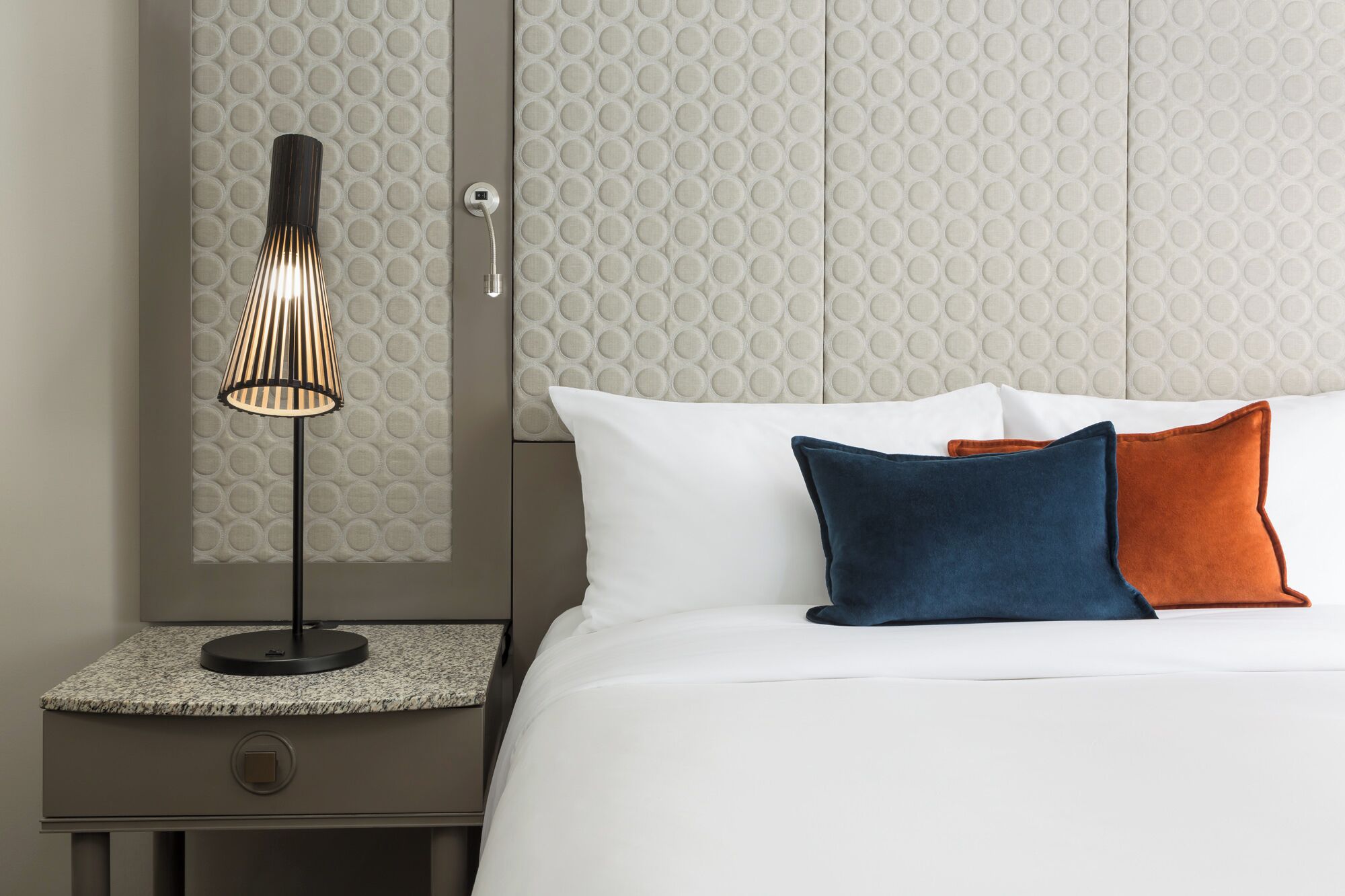 Ein ordentlich bezogenes Bett mit weißen und zwei farbigen Kissen, eine moderne Lampe auf einem Nachttisch und eine strukturierte Wand dahinter.