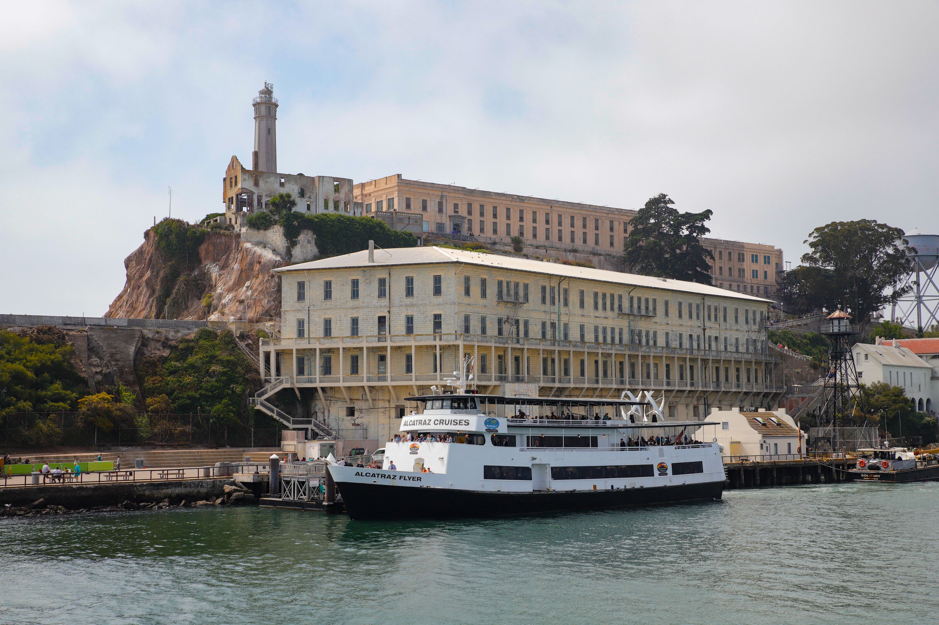 A large white cruise ship docked at a pier with a historic building and lighthouse in the background.