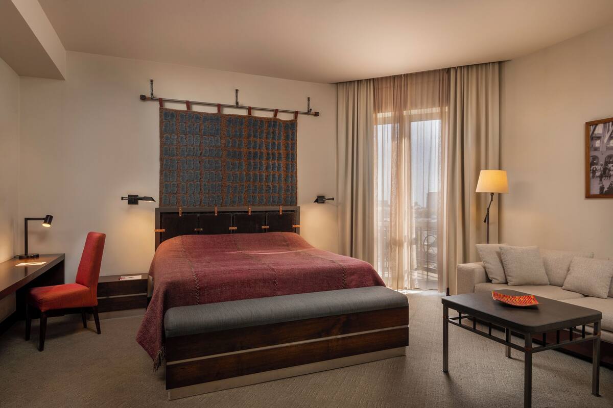 Ein Hotelzimmer mit Bett, Schreibtisch, Sessel, Schlafsofa, Lampe und einem fenster mit Vorhängen.