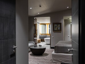 A modern hotel room with a large bed, a round coffee table, a sofa, and a view into the bathroom.